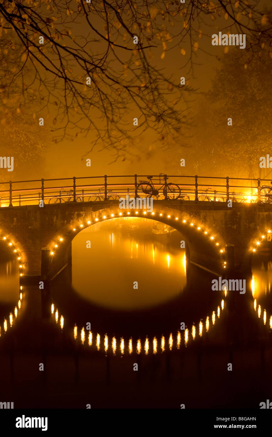 Ponte illuminato riflessa nel canale in una nebbiosa notte a Amsterdam Paesi Bassi Foto Stock