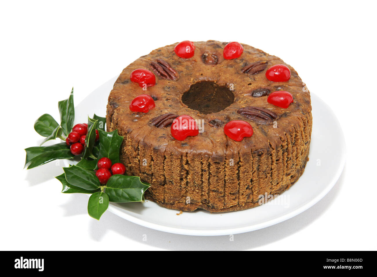 Bella torta di frutta di Natale guarnita con ciliegie di noci pecan e un rametto di agrifoglio isolati su sfondo bianco Foto Stock