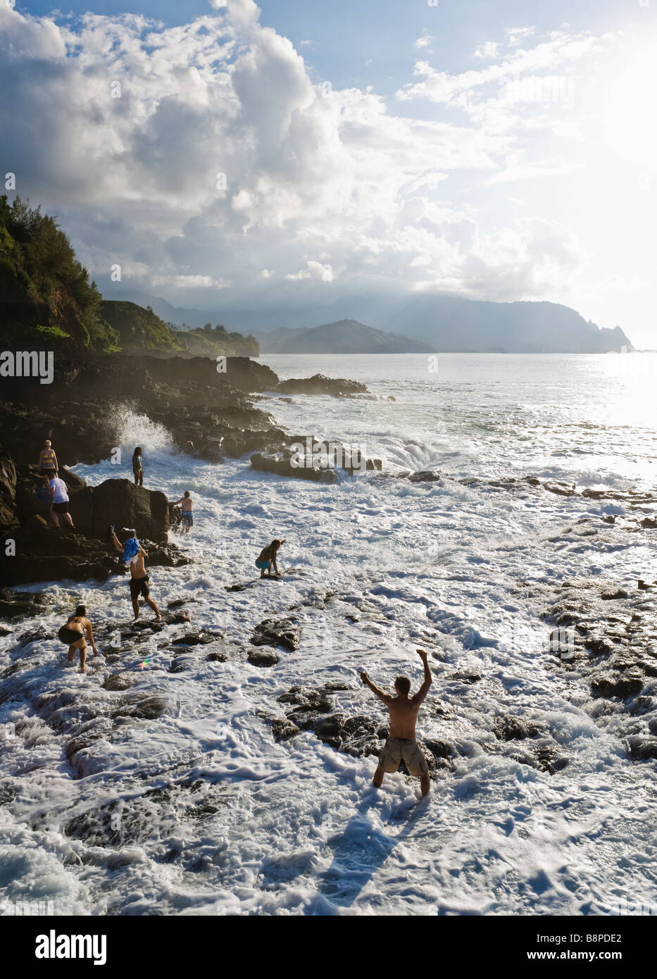 I turisti mantenendo la costa rocciosa come una sorpresa grande onda lavaggi completamente al di sopra del bagno di Queens nei pressi di Princeville, Kauai, HA Foto Stock