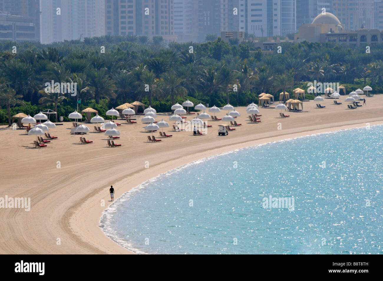 Dubai vicino a Palm Island spiaggia privata di sabbia artificiale curva sole Sedie a sdraio ombrelloni e vegetazione ombreggiata edifici alti oltre United Arab Emirati Arabi Uniti Foto Stock