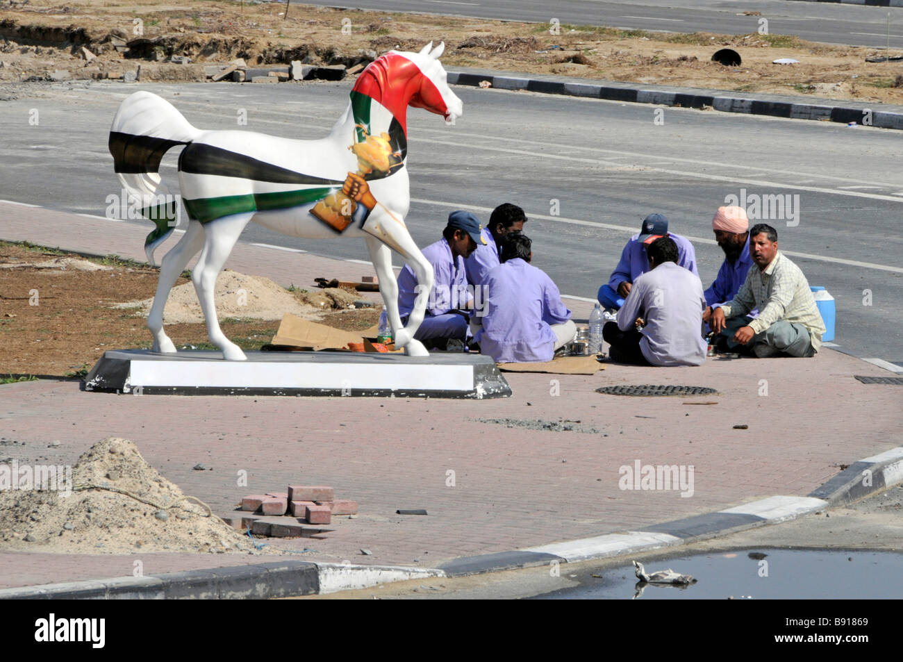 Dubai strada scena fuori edificio cantiere gruppo di lavoratori Durante la pausa pasto seduto sul marciapiede con statua a cavallo unita Emirati Arabi Uniti Foto Stock