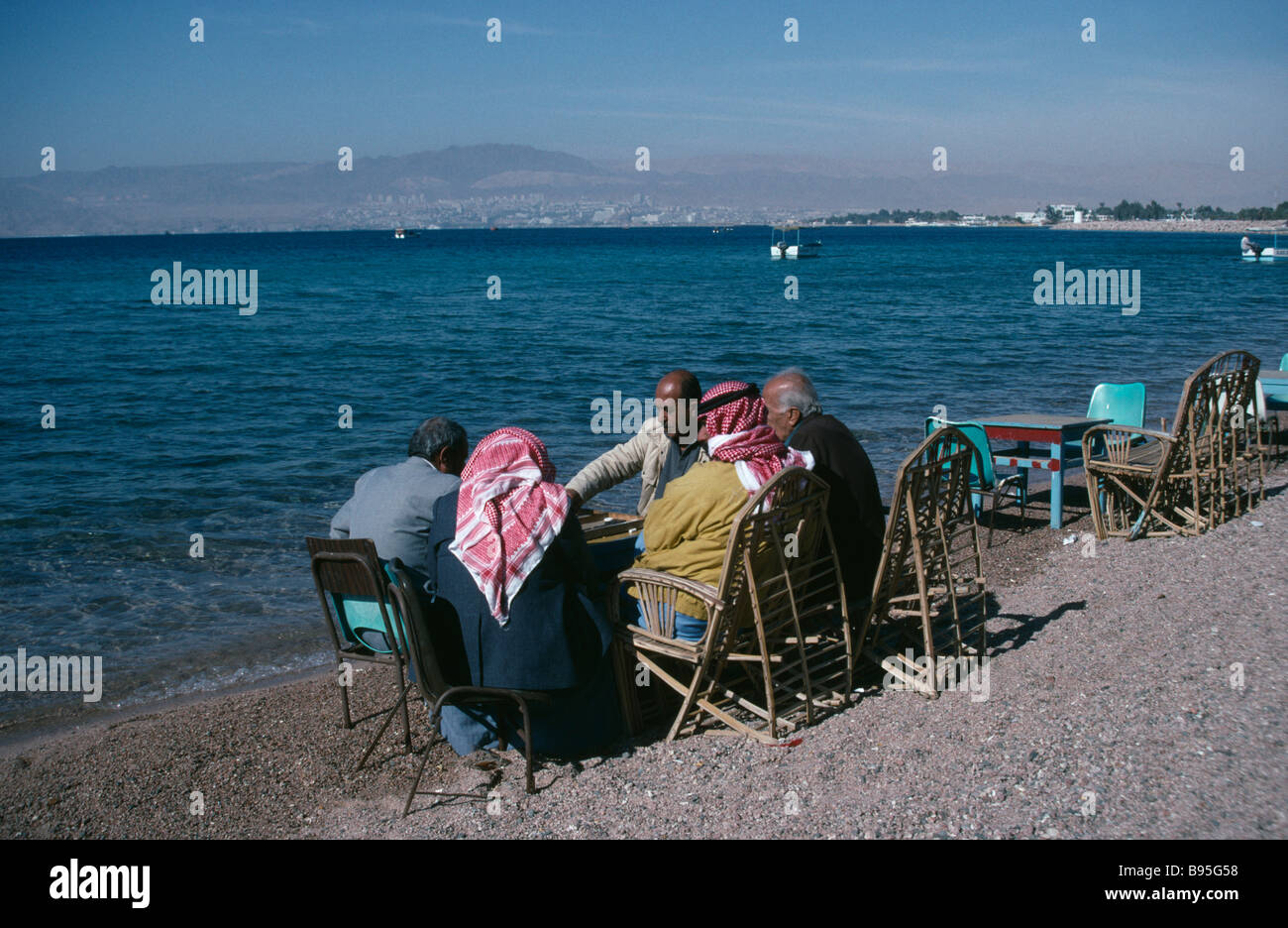 La Giordania Aqaba gruppo di uomini di giocare a backgammon sulla riva della spiaggia con Eliat e Israele a distanza dietro. Foto Stock