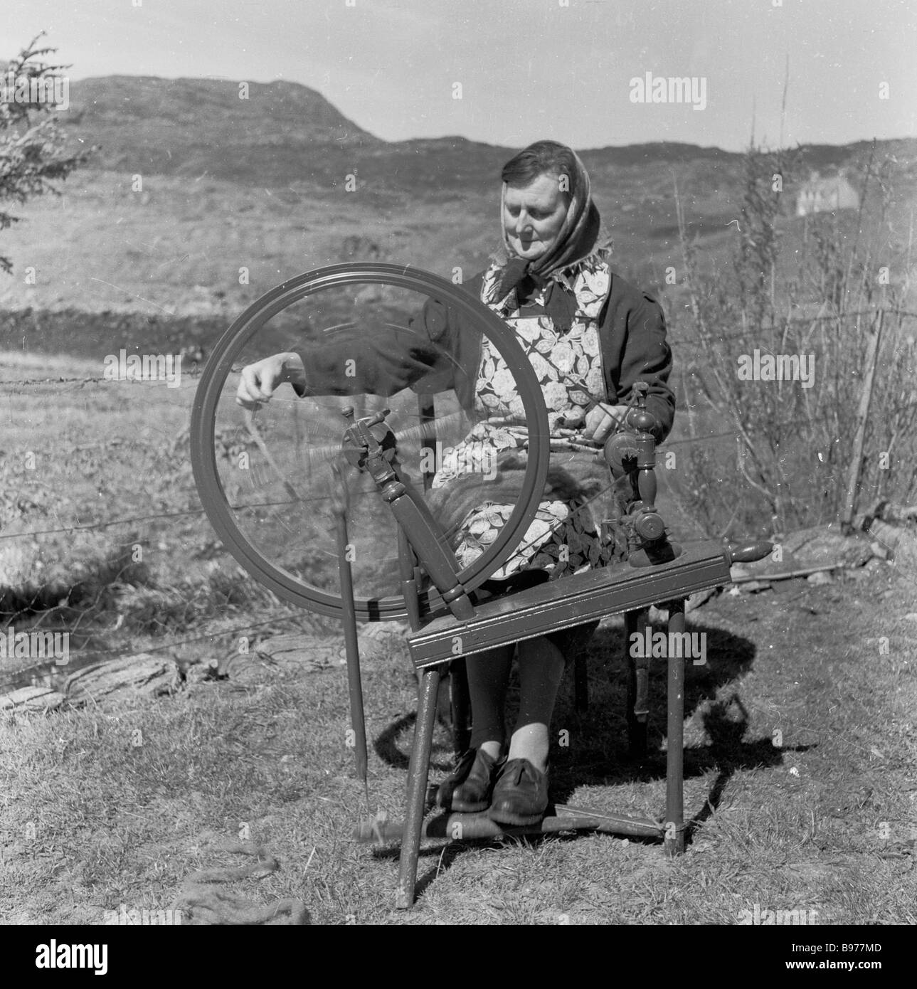 1950s, storica, un'anziana signora scozzese crofter seduto a lavorare una ruota di legno che gira la lana fuori sul suo croft, Highlands, Scozia, Regno Unito Foto Stock