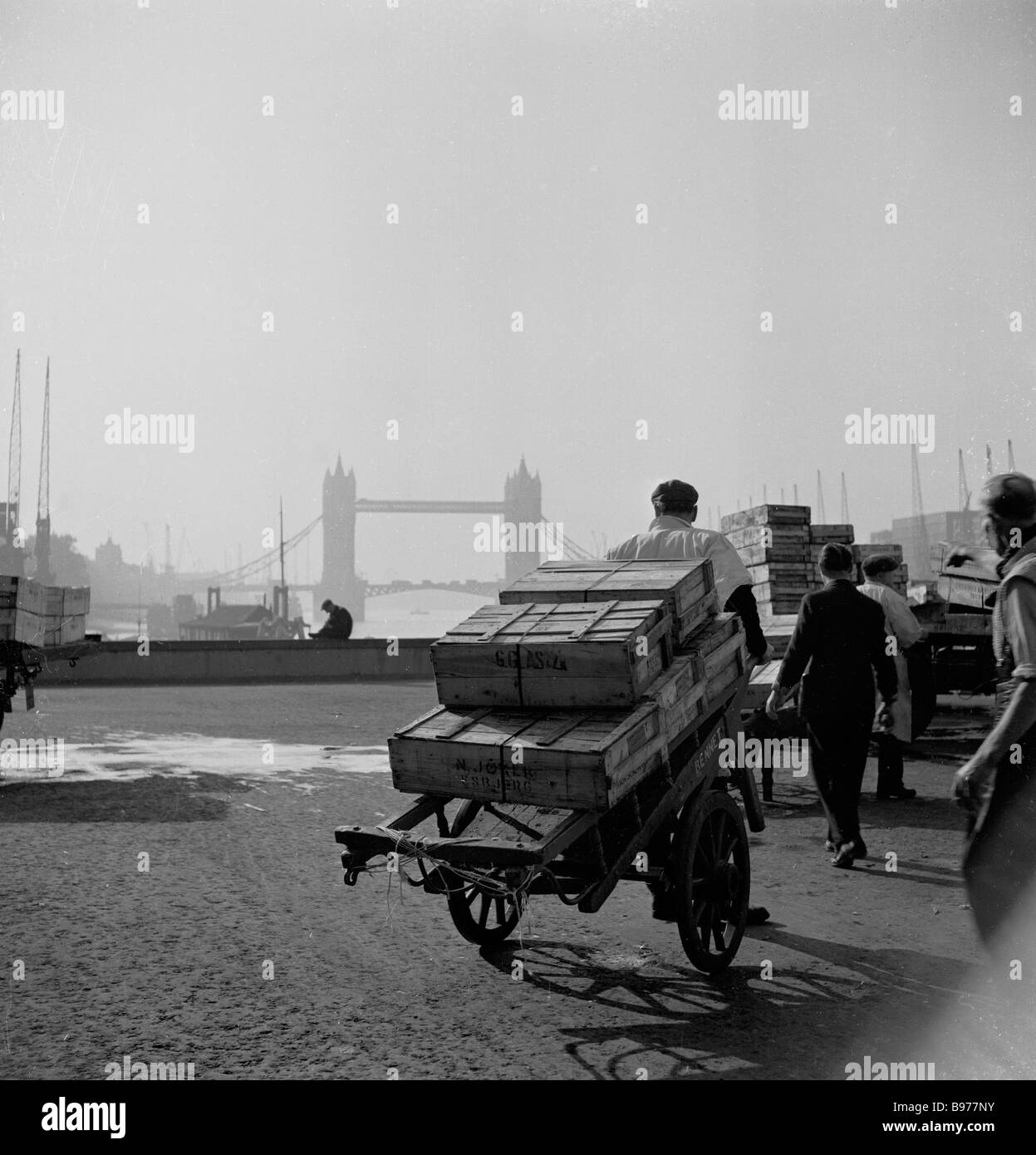 1950s, storici, i portieri del mercato tirano i loro carri attraverso lo storico London Bridge, Londra, Inghilterra. Il famoso Tower Bridge è visto in lontananza. Foto Stock