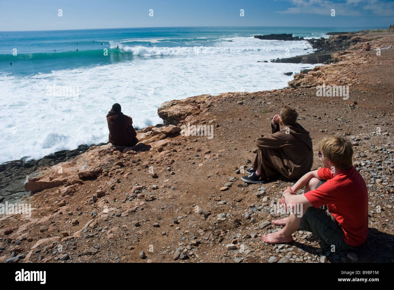 Tre surfers sedersi sul punto in caldaie surf break e guardare il rigonfia rotolare giù per le scogliere. Foto Stock