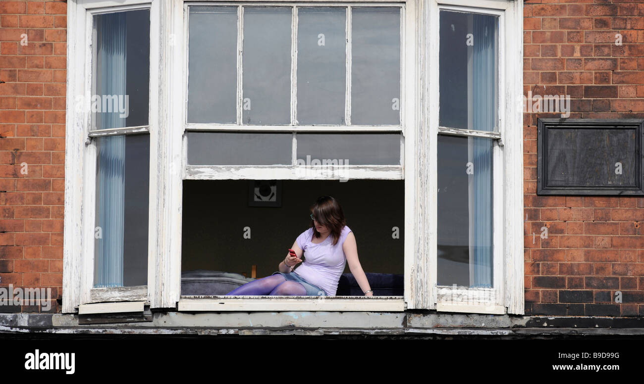 Una ragazza reclinabile in un appartamento al di sopra di un negozio, inviare un messaggio di testo sul suo telefono cellulare da una finestra aperta. Foto Stock