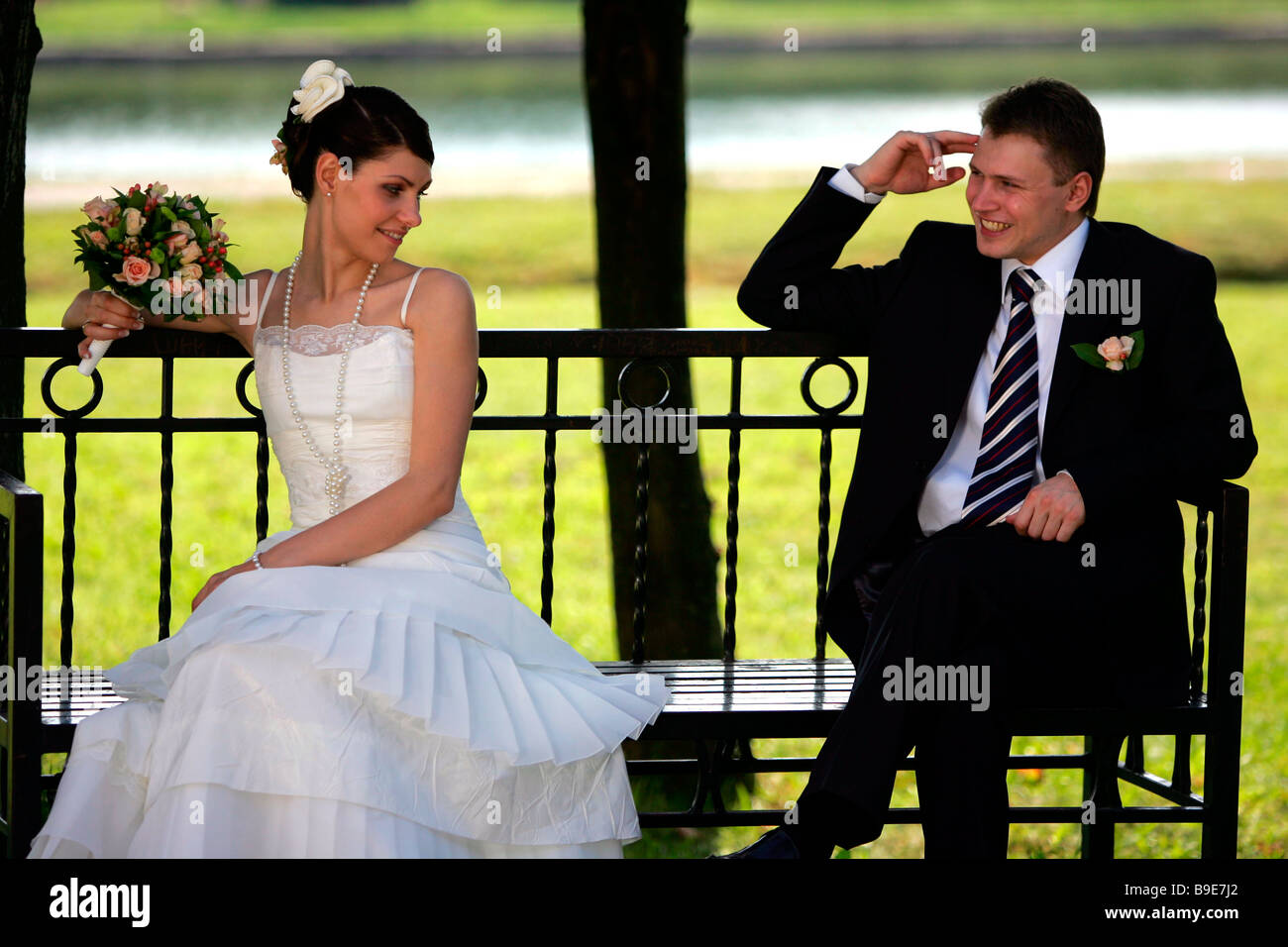 Sposa giovane seduto su una panchina nel parco sorridente groom e coy sposa con bouquet Foto Stock