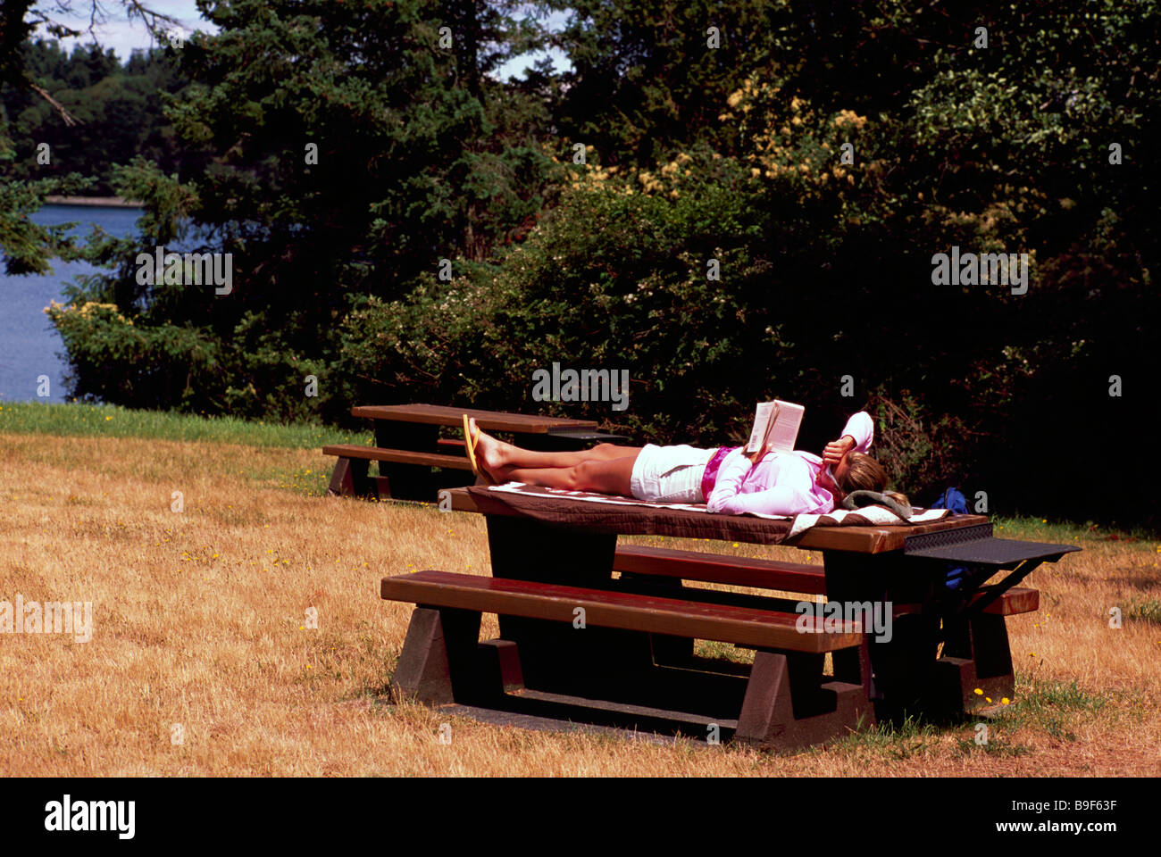 Donna sdraiata in Sun sulla parte superiore del tavolo da picnic e la lettura di un libro Foto Stock