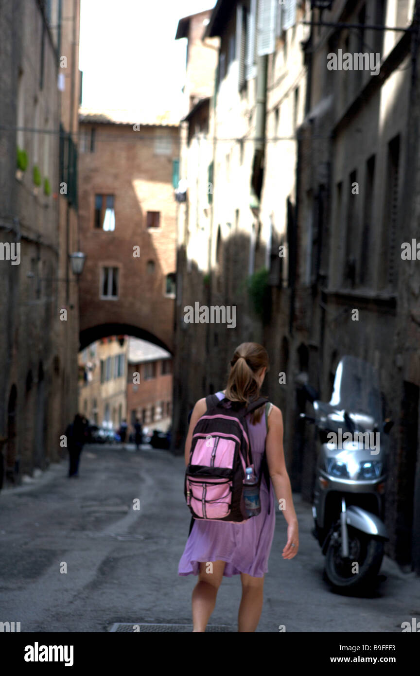 Una giovane ragazza con una rosa zaino camminate giù per una strada laterale a Siena Italia e verso uno scooter con un arco a distanza Foto Stock