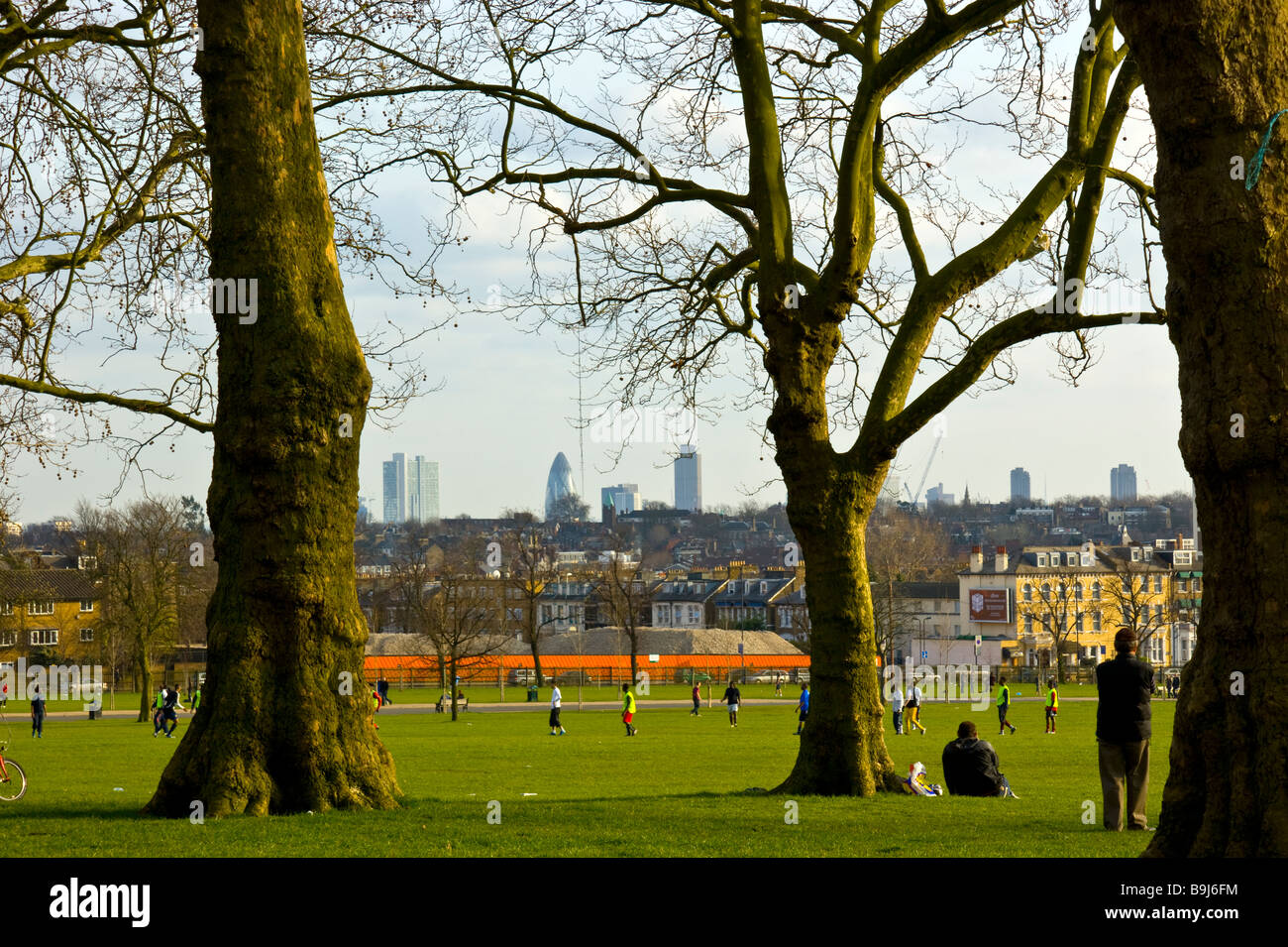 Domenica pomeriggio a Finsbury Park. Coppia alberi di quercia e gli spettatori in occasione di una partita di calcio con la City of London skyline a distanza Foto Stock