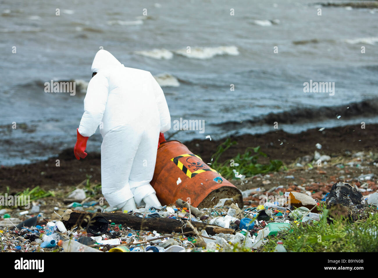 Persona in tuta protettiva che trasportano barile di rifiuti pericolosi sulla riva inquinati Foto Stock