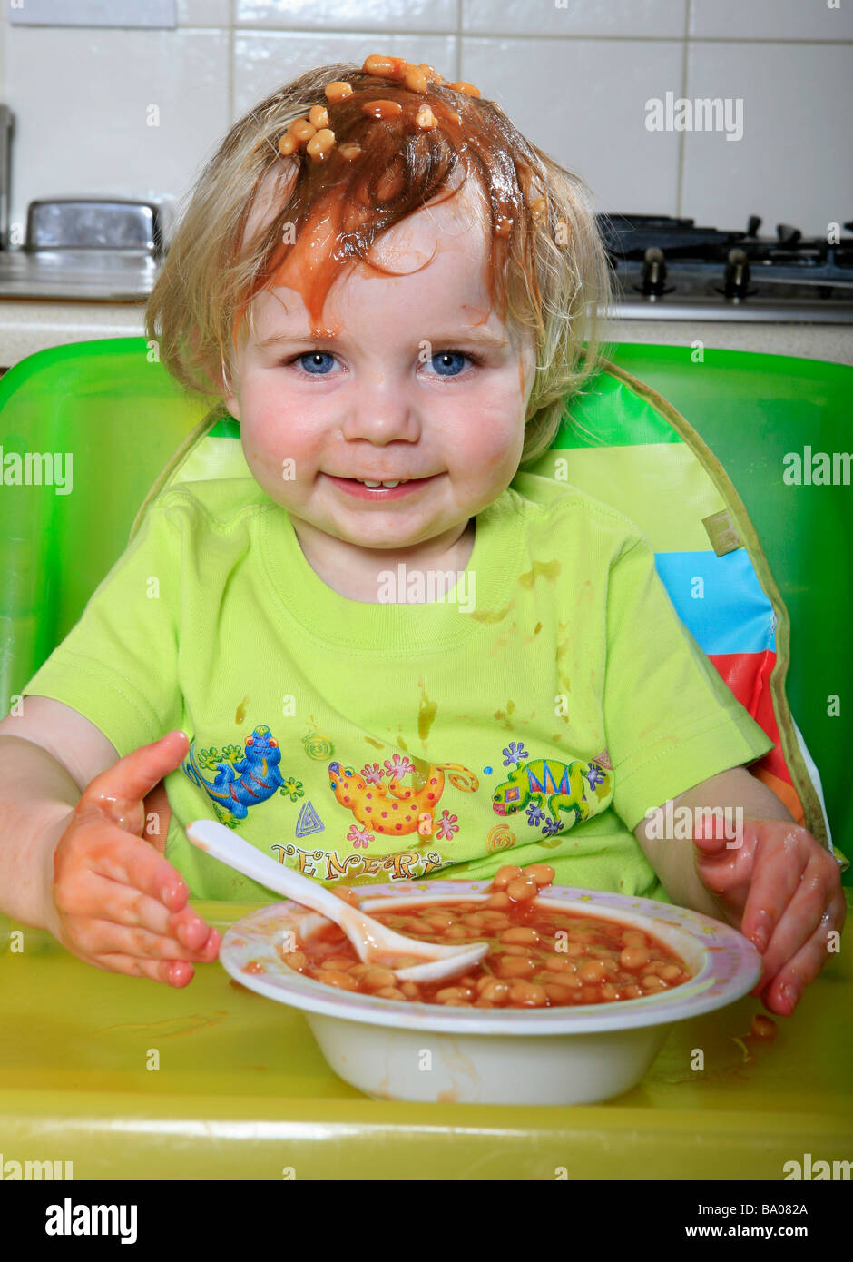 Bambino con fagioli al forno sulla testa sorridente. bambino mangiare i fagioli il bambino nel seggiolino bambino in cucina Foto Stock