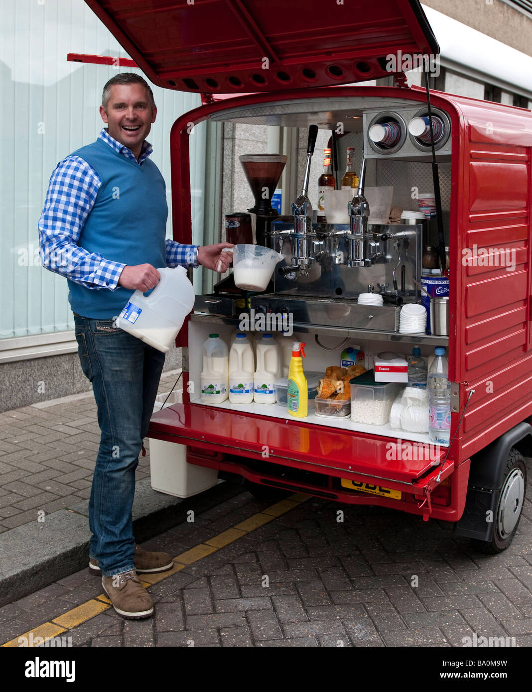 Cheeky , vivace , felice , sorridente venditore ambulante commerciante di mercato la vendita di bevande calde dal suo cellulare, macchina per il caffè è un furgone. Foto Stock