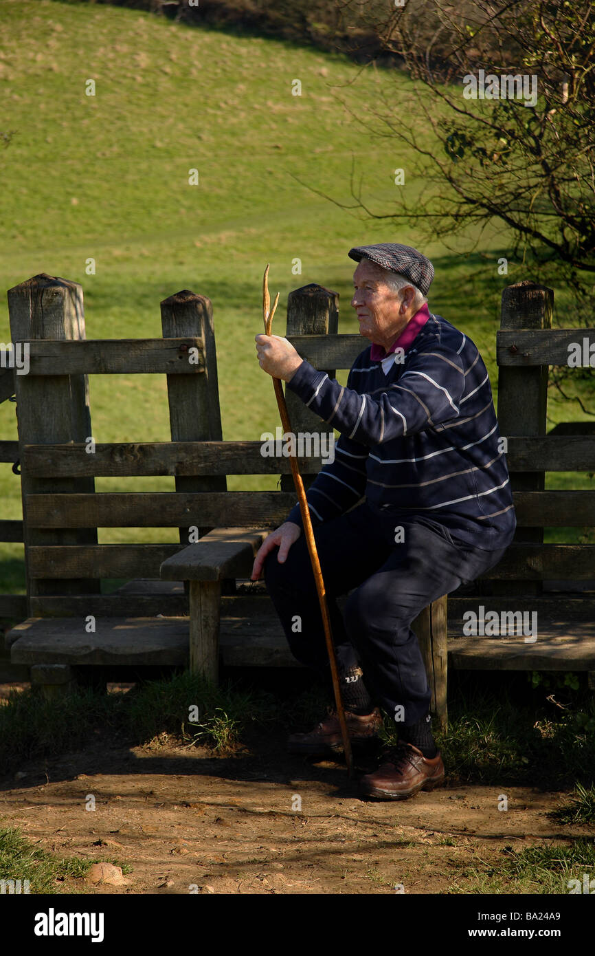 Un vecchio uomo con un alto bastone seduto su uno stile vicino a Golden cap in appoggio giorno soleggiato Campo dietro il Dorset Foto Stock