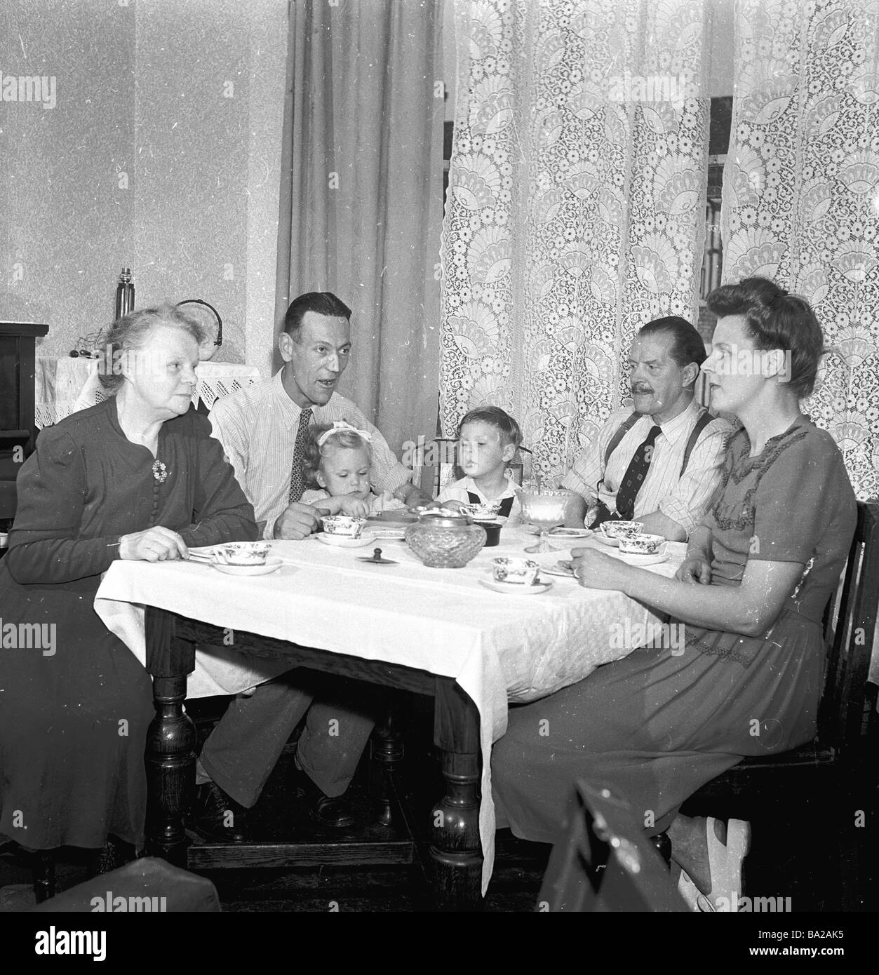1950s, storico, vita familiare in Gran Bretagna. Generazioni diverse si siedono intorno ad un piccolo tavolo di legno coperto di stoffa che ha il tè insieme. Foto Stock