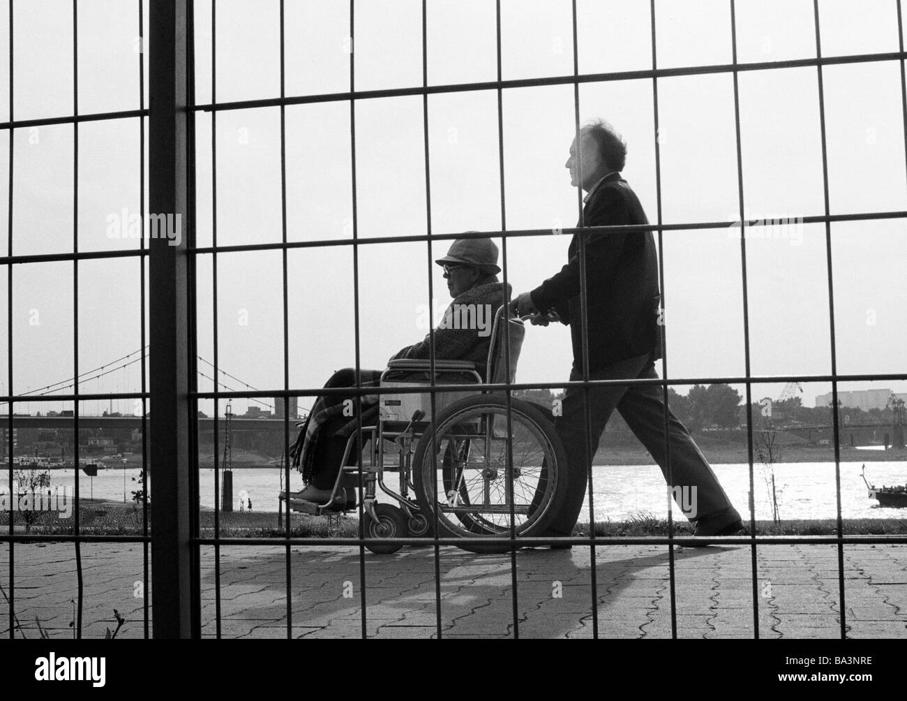 Negli anni settanta, foto in bianco e nero, persone di handicap fisici, aiuti, uomo anziano in sedia a rotelle sarà spinto da un giovane uomo, vista attraverso un reticolo recinto, di età compresa tra 70 a 80 anni di età compresa tra 40 a 50 anni Foto Stock