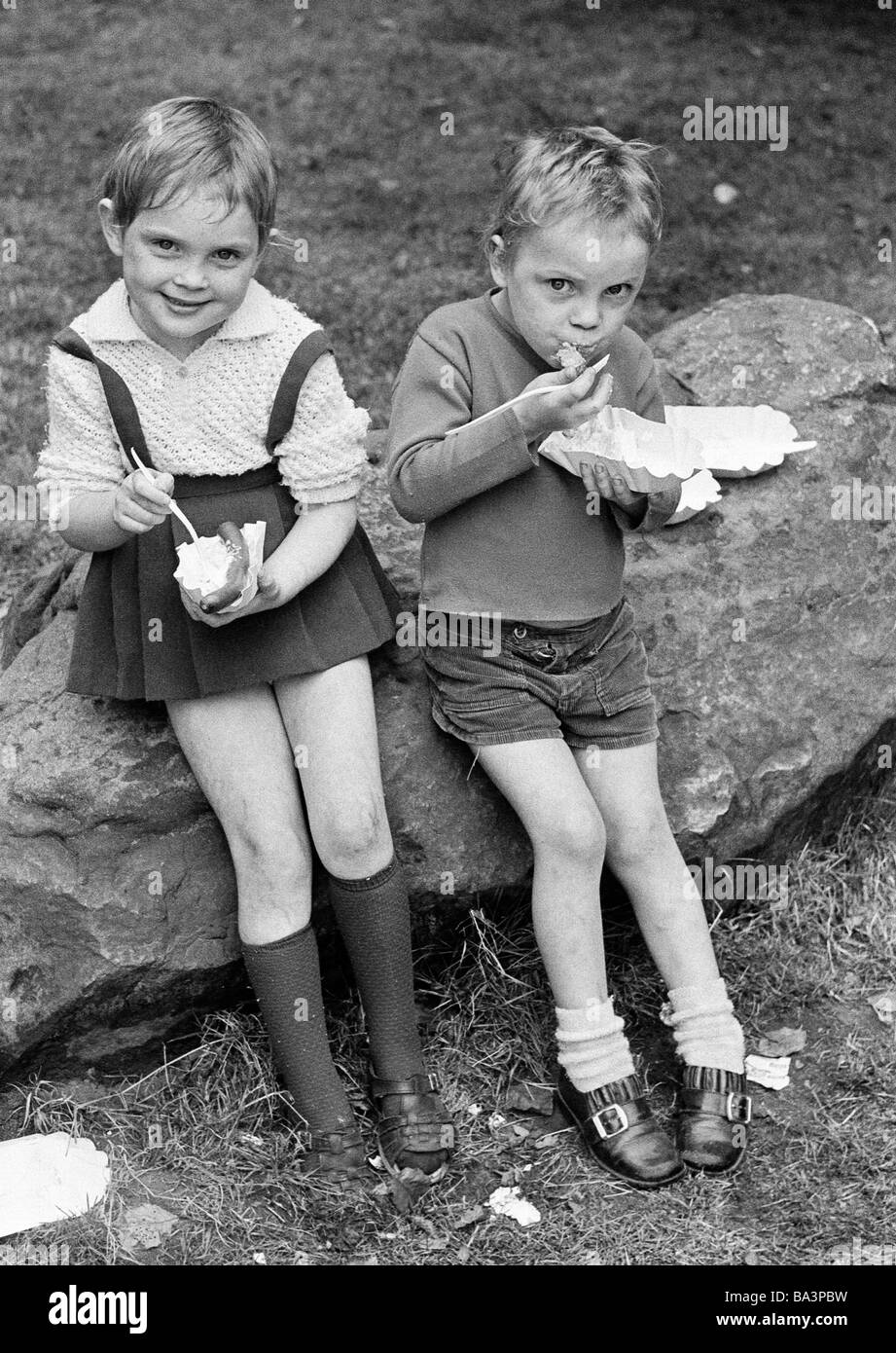 Negli anni settanta, foto in bianco e nero, persone, bambini, bambina e little boy seduti fianco a fianco su una roccia e mangiare un fast food, salsiccia e insalata di patate, di età compresa tra i 4 ed i 7 anni Foto Stock