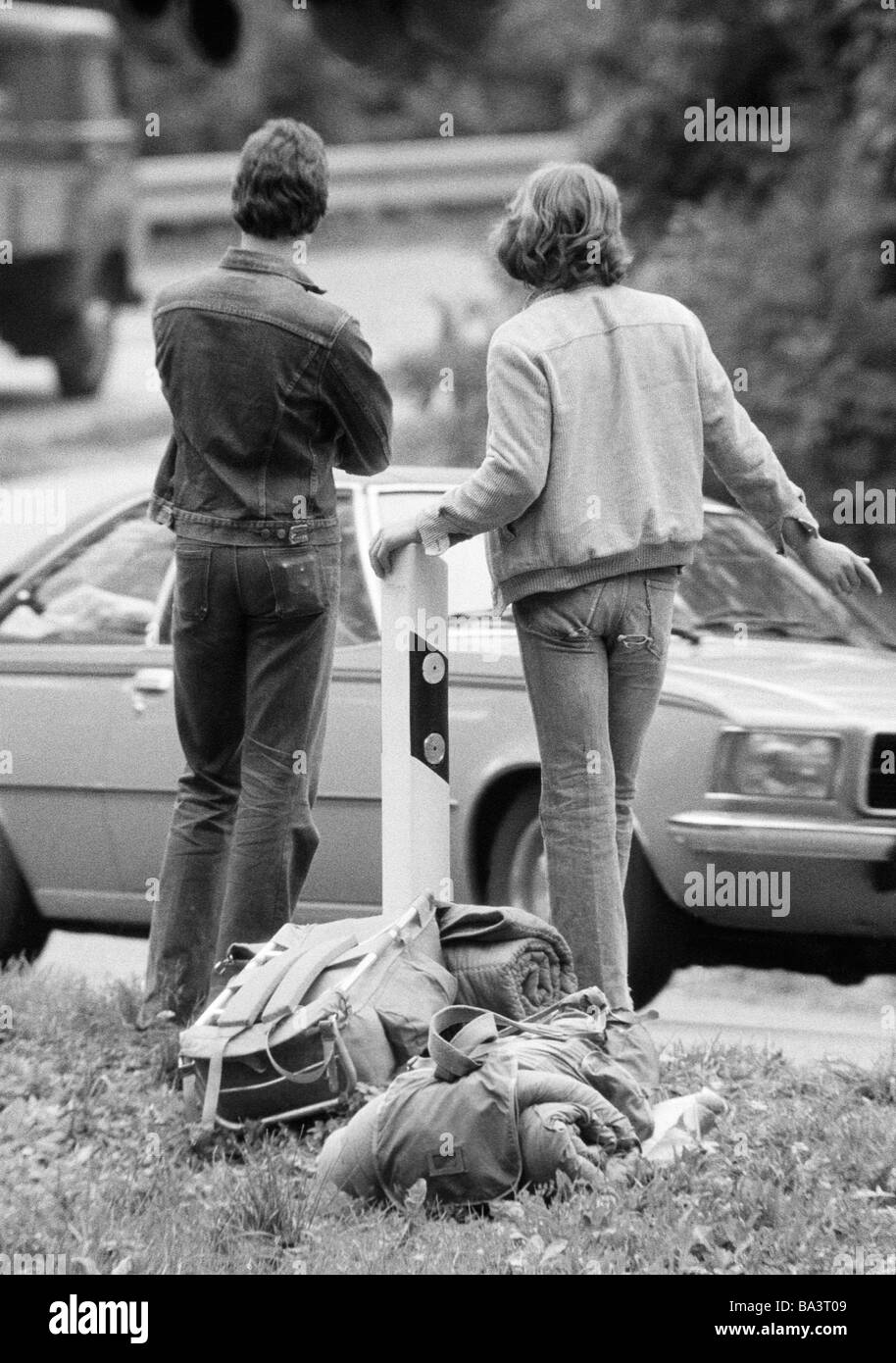 Negli anni settanta, foto in bianco e nero, persone, due giovani hitchhikers in piedi in una strada, bagagli, vista posteriore, una macchina passa dai 16 ai 20 anni Foto Stock
