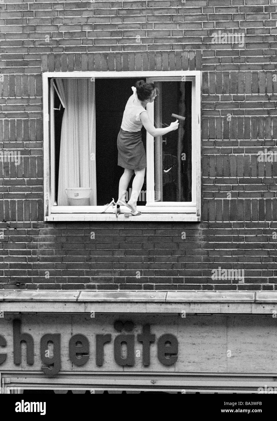Negli anni settanta, foto in bianco e nero, persone, pericolo di incidente, giovane casa-moglie indossando muli pulisce una finestra al piano superiore di una casa e si piega in avanti, una gamba sorge sul davanzale esterno, di età compresa tra 25 a 35 anni Foto Stock