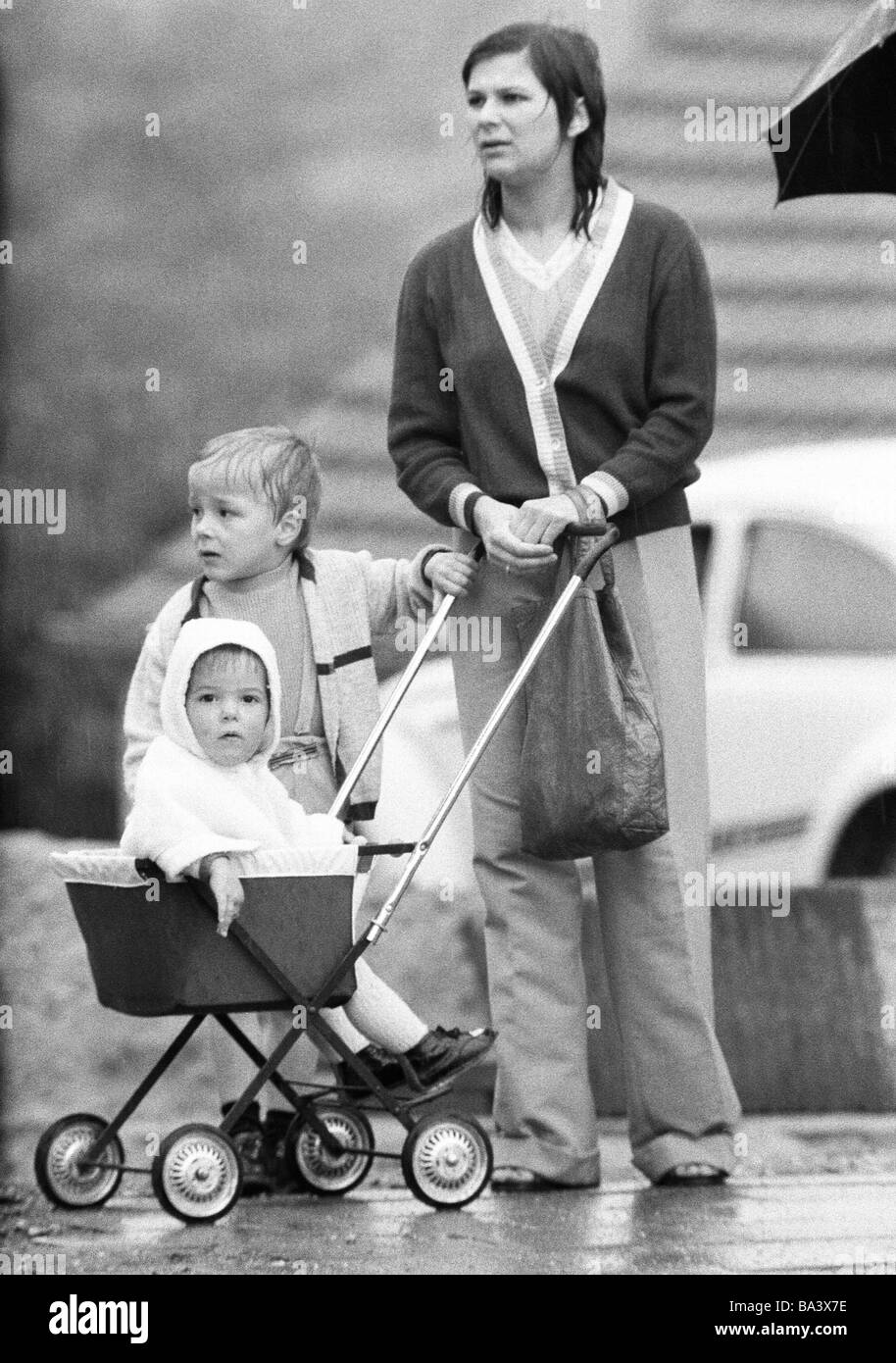 Negli anni settanta, foto in bianco e nero, persone, madre e due figli con un bambino carrello attendere sotto la pioggia, donna di età compresa tra i 30 e i 35 anni, ragazzo di età compresa tra 3 e 4 anni, ragazza, di età compresa tra 1 e 2 anni Foto Stock