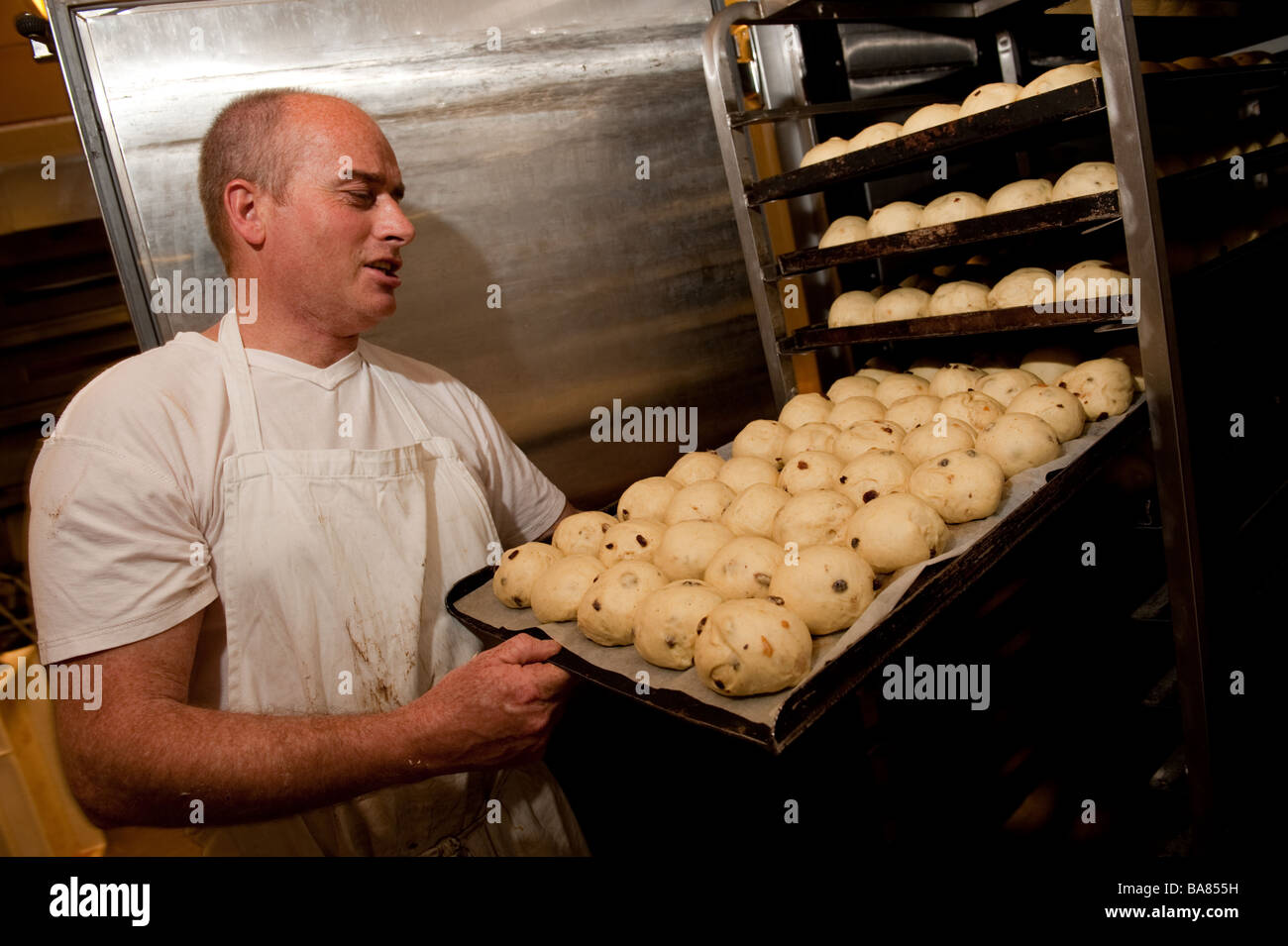 Un baker preparare calde tradizionali ciambelle trasversale per la pasqua UK - prendendo le ciambelle sollevata al di fuori dell'armadio di riscaldamento prima della cottura Foto Stock