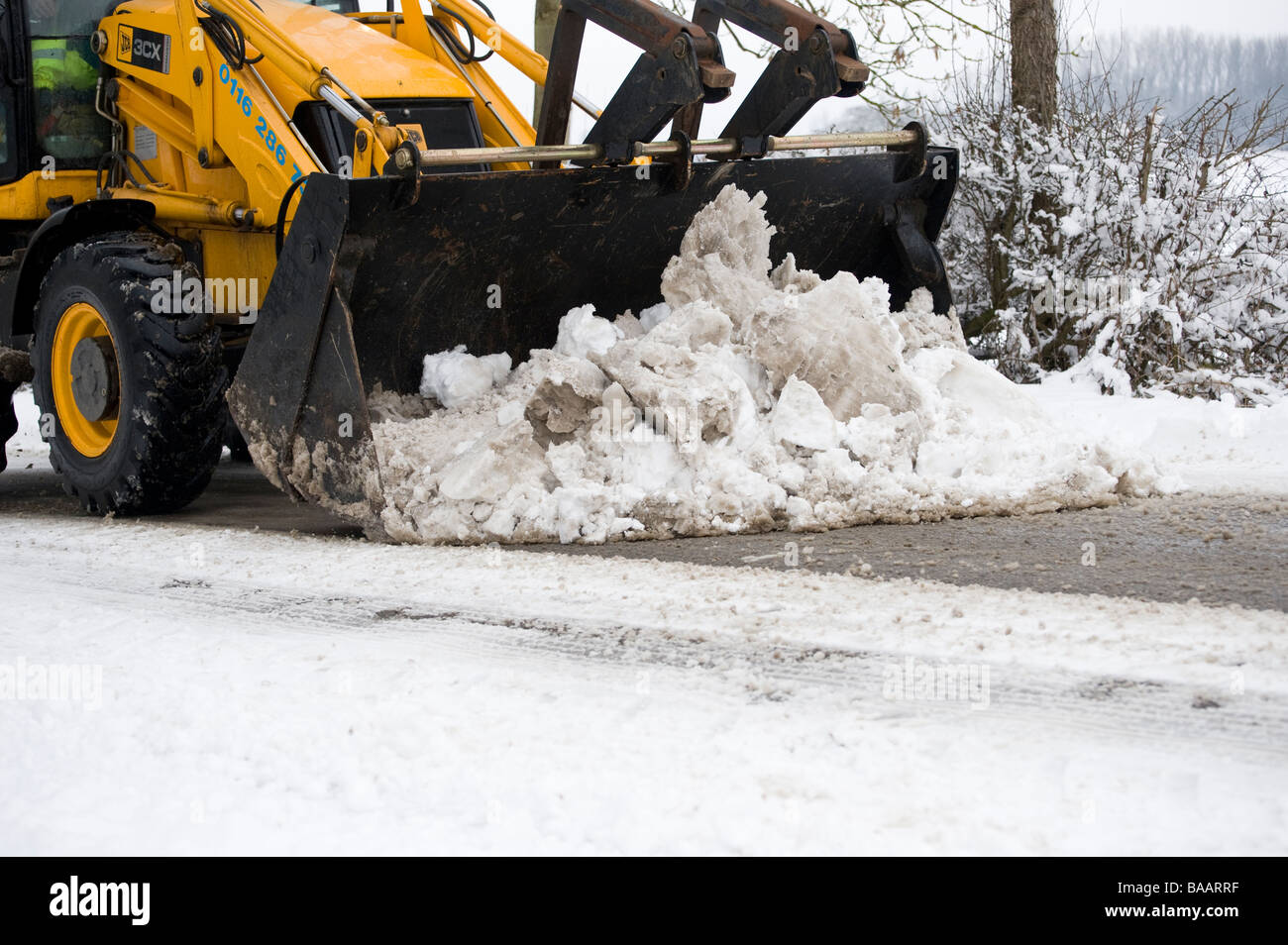 JCB con benna pala essendo utilizzato per cancellare una coperta di neve strada di campagna in Inghilterra al tempo di inverno Foto Stock