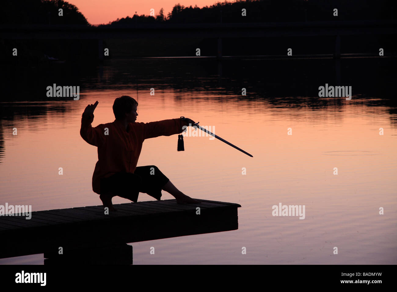 Una giovane donna in silhouette su un molo al tramonto praticare il tai chi con una spada Limousin Francia Foto Stock