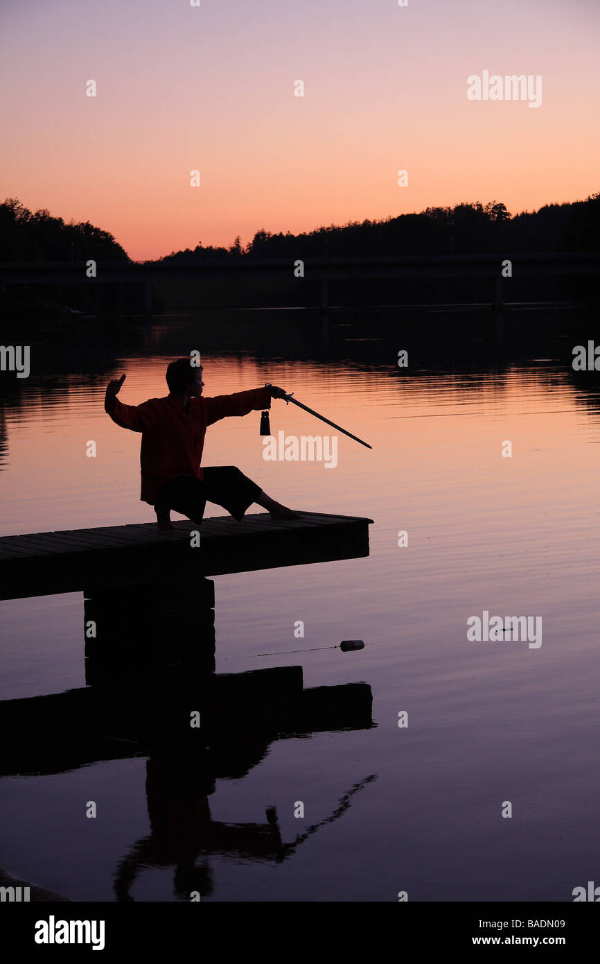Una giovane donna in silhouette su un molo al tramonto praticare il tai chi con una spada Limousin Francia Foto Stock