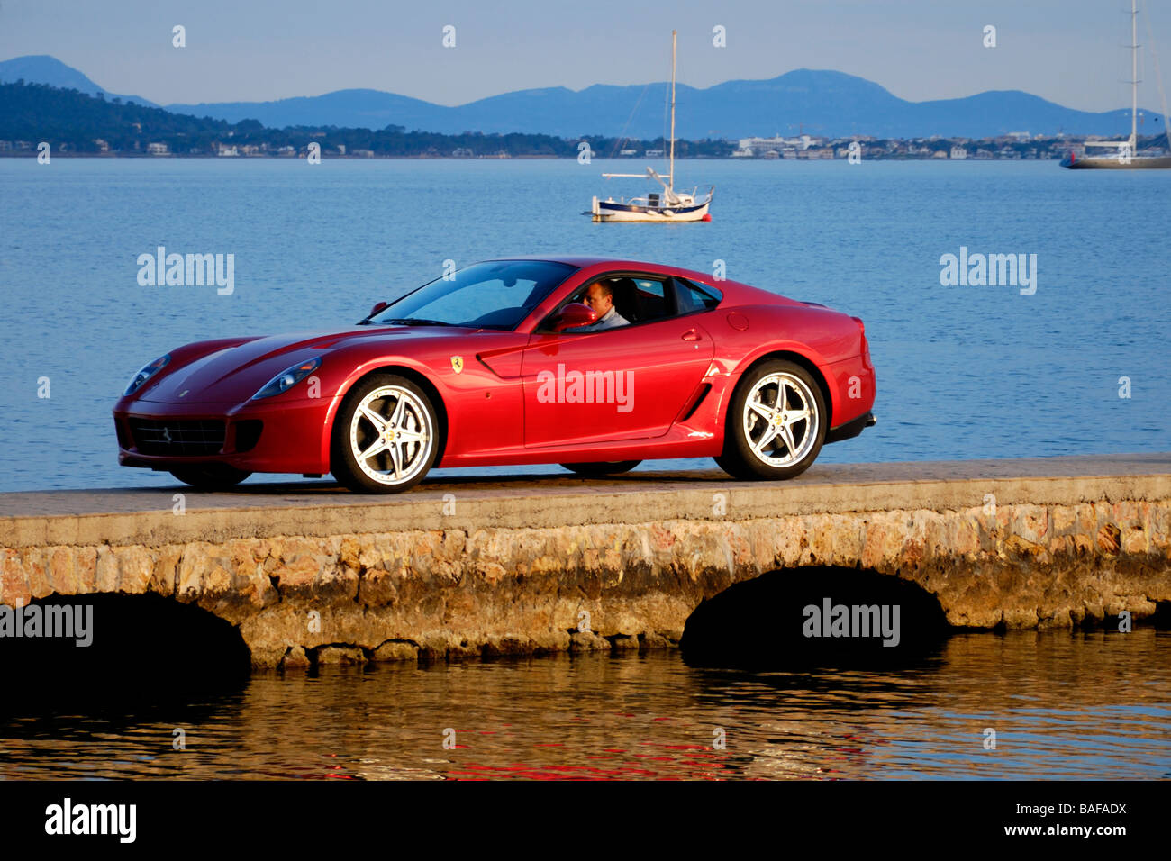 Rosso Ferrari con autista privato sulla dock mediterranea a Pollensa bay con yacht e delle montagne sullo sfondo al tramonto Foto Stock