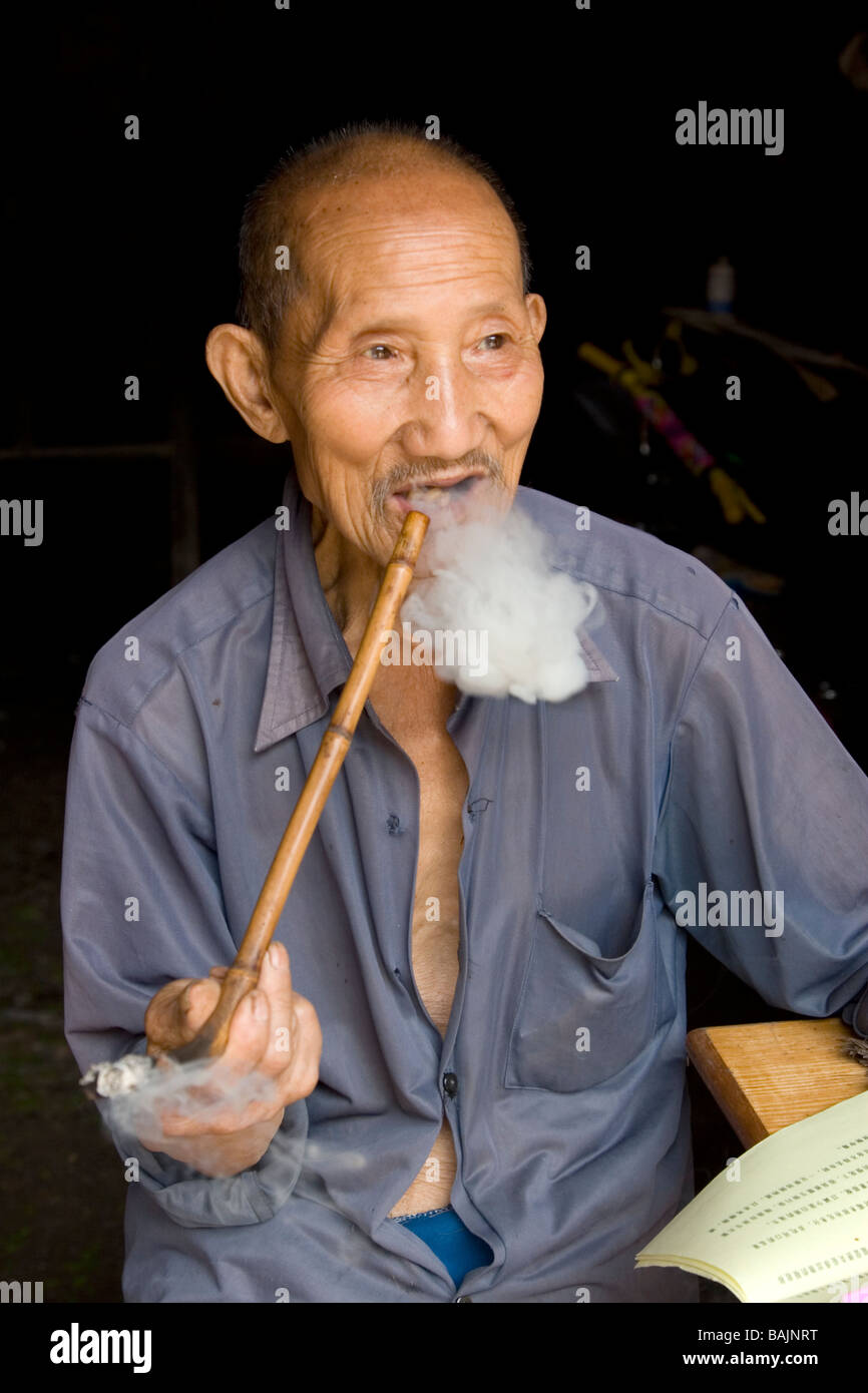 Un vecchio uomo nel cortile della sua casa colonica di fumare fatti in casa a base di tubo e la lettura oroscopo cinese materiale nel piccolo villaggio. Foto Stock