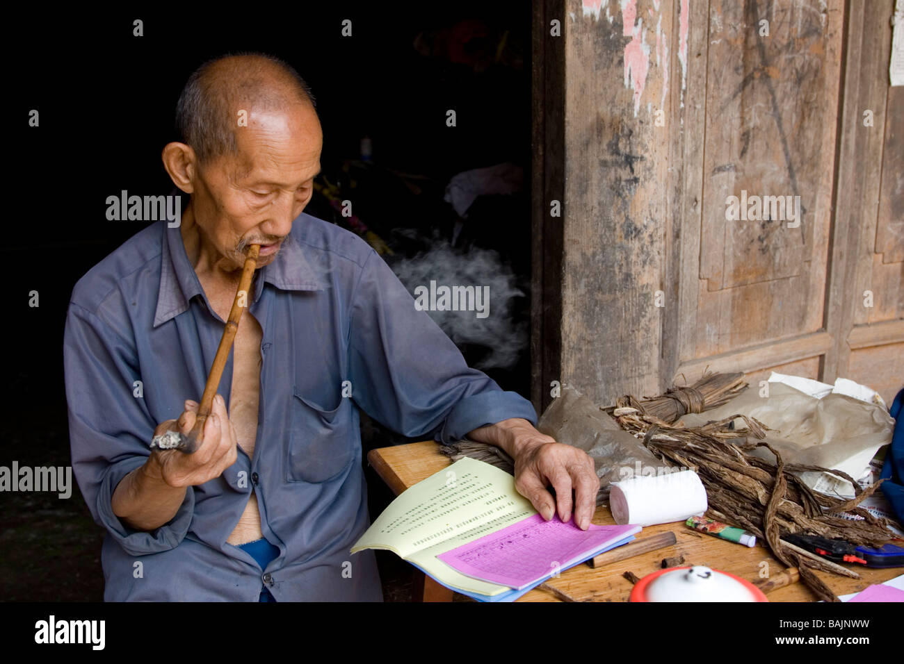 Un vecchio uomo nel cortile della sua casa colonica di fumare fatti in casa a base di tubo e la lettura oroscopo cinese materiale nel piccolo villaggio. Foto Stock