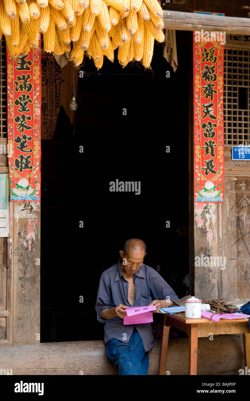 Un vecchio uomo nel cortile della sua casa colonica di fumare fatti in casa a base di tubo e la lettura oroscopo cinese materiale nel piccolo villaggio. Foto Stock