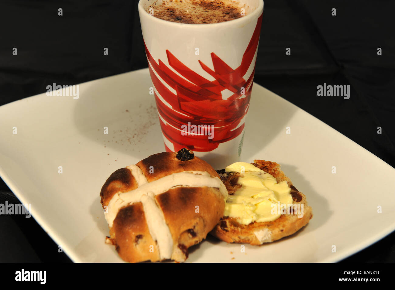 Tostato e imburrato hot cross bun e cappuccino serviti su un tè bianco e piastra di sfondo nero. Foto Stock
