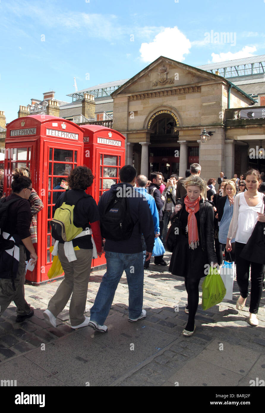 La folla di turisti e amanti dello shopping a piedi passato cabine telefoniche rosse sotto il sole al di fuori del mercato di Covent Garden di Londra. Foto Stock
