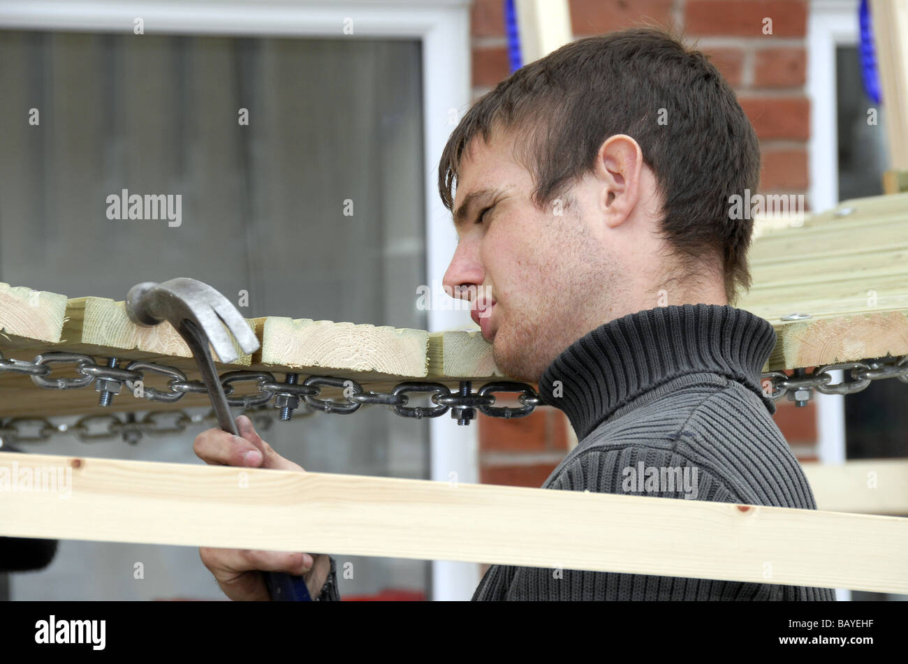 Giovane maschio builder azienda martello occhiatura-up la linea di vista sulla catena-ponte di collegamento sul legname climbing frame progetto di fabbricazione. Foto Stock