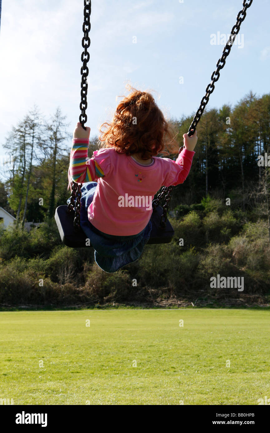 Bambina di tre anni con i capelli rossi su altalena nel parco in una giornata di sole. Foto Stock