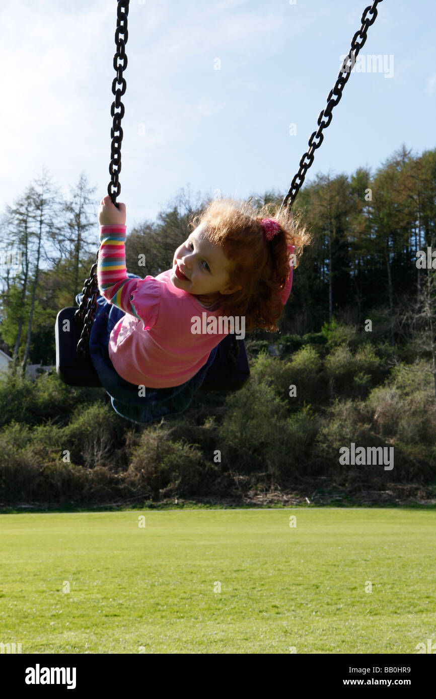 Bambina di tre anni con i capelli rossi su altalena nel parco in una giornata di sole. Foto Stock