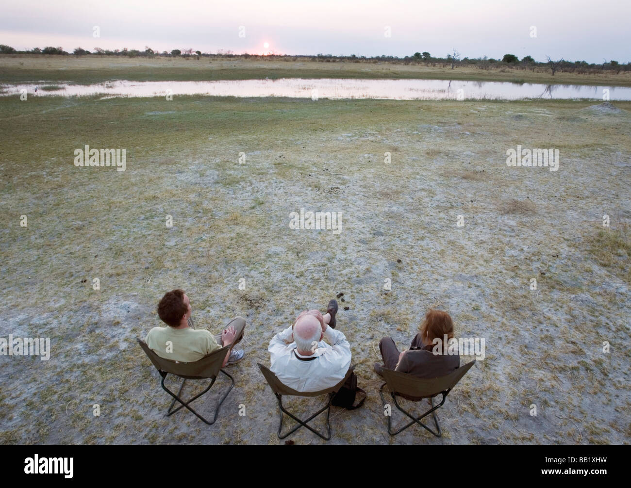 Ampio angolo di visione di tre persone su safari a guardare il sole che tramonta, Okavango Delta, Botswana Foto Stock