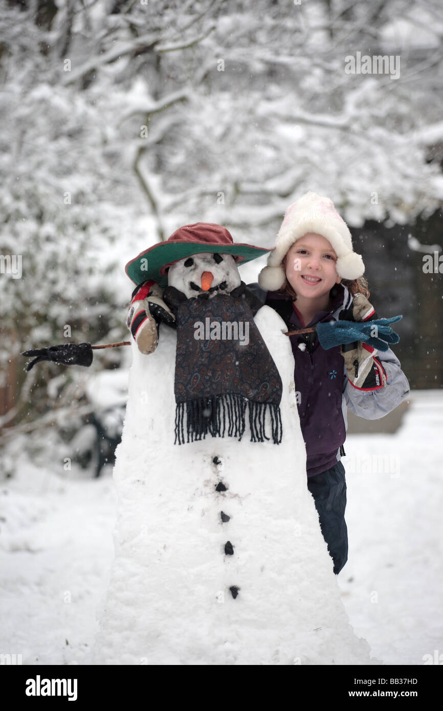 5 anno vecchia ragazza orgogliosamente sorge accanto al suo pupazzo di neve nel giardino di casa sua. Foto Stock