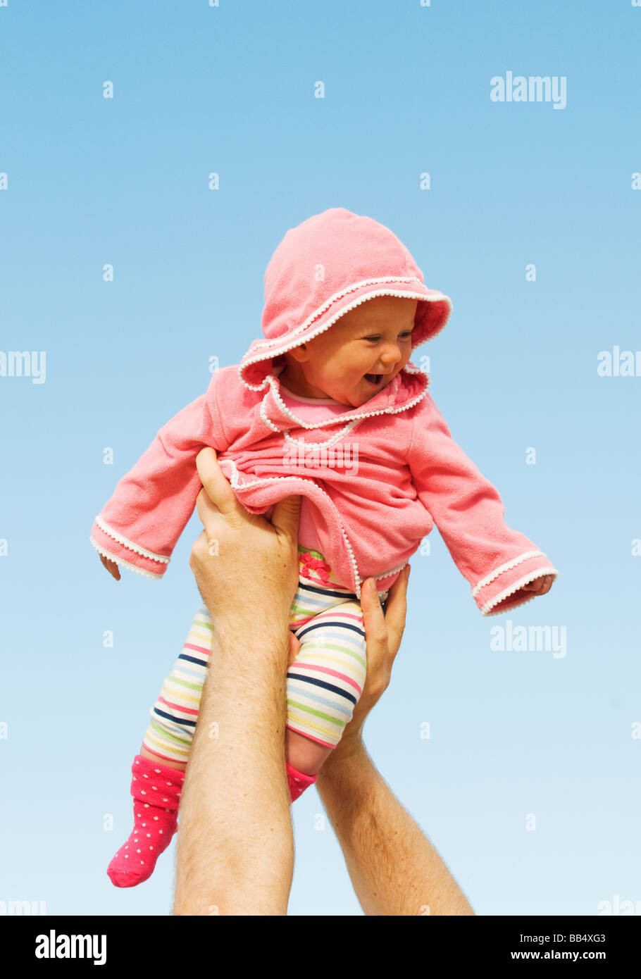 BABY GIRL, 9 mesi di età, sorridendo felice, tenuto alto in aria da bracci di adulto contro il cielo blu. Foto Stock