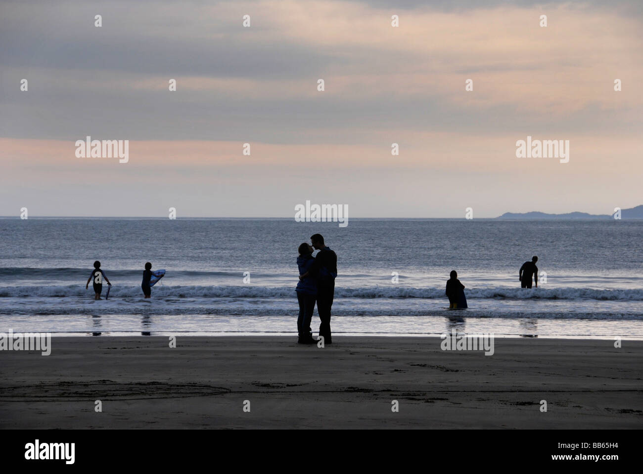 Giovane kiss in silhouette sulla spiaggia in serata in Galles mentre i bambini surf Foto Stock