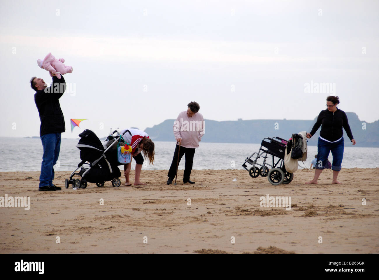 L'uomo detiene baby fino in aria sulla spiaggia mentre le donne in lotta con i passeggini Foto Stock