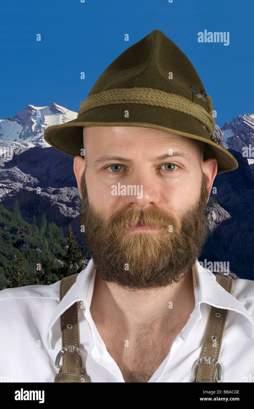 Un uomo che indossa un cappello alpino e lederhosen con uno sfondo montano Foto Stock