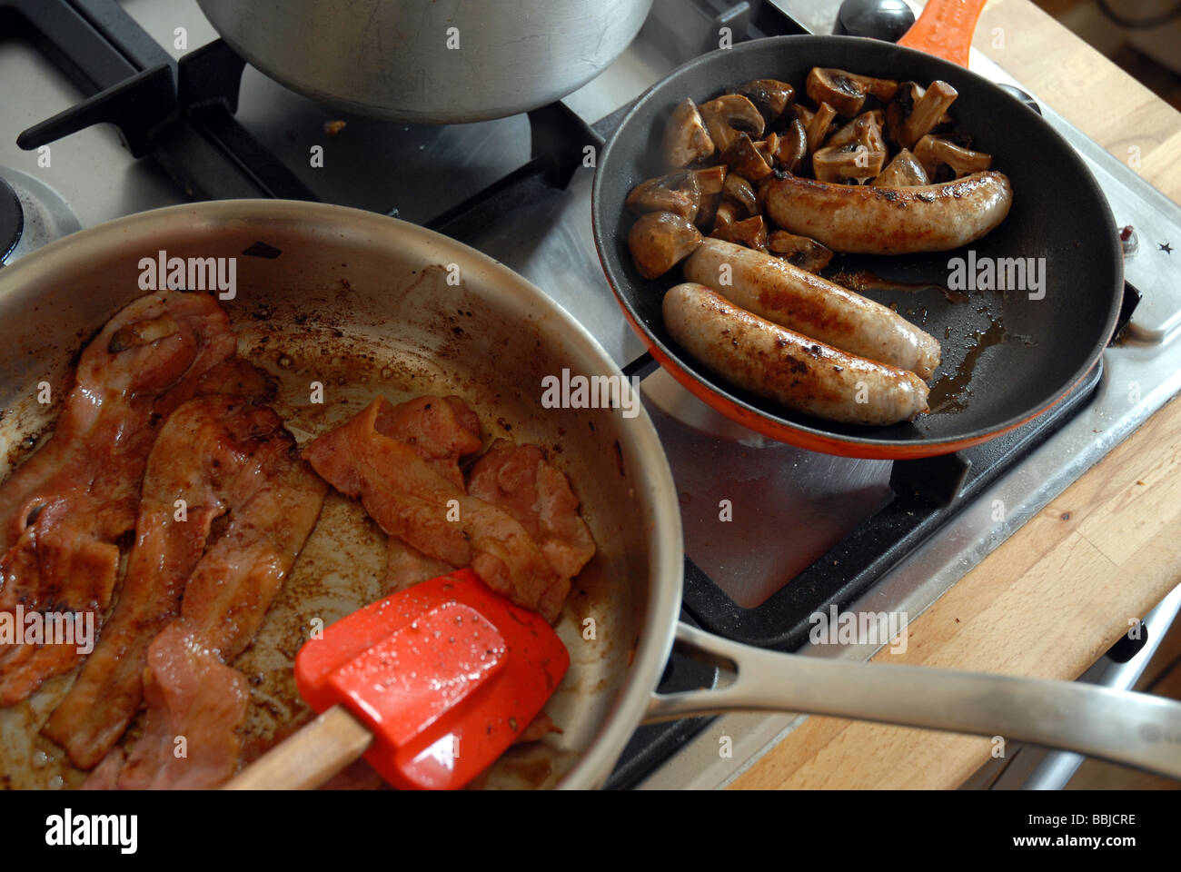 Salsicce e pancetta fritte in padelle su piano cottura a gas per una colazione all'inglese completa tradizionale Foto Stock