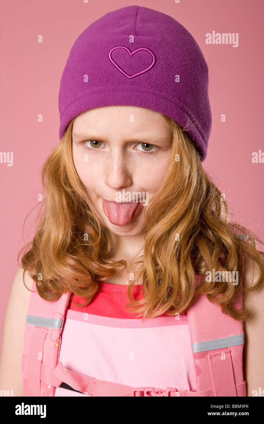 I Capelli rossi ragazza indossando un viola cofano e zainetto davanti a uno sfondo di colore rosa, prendendo la sua lingua Foto Stock