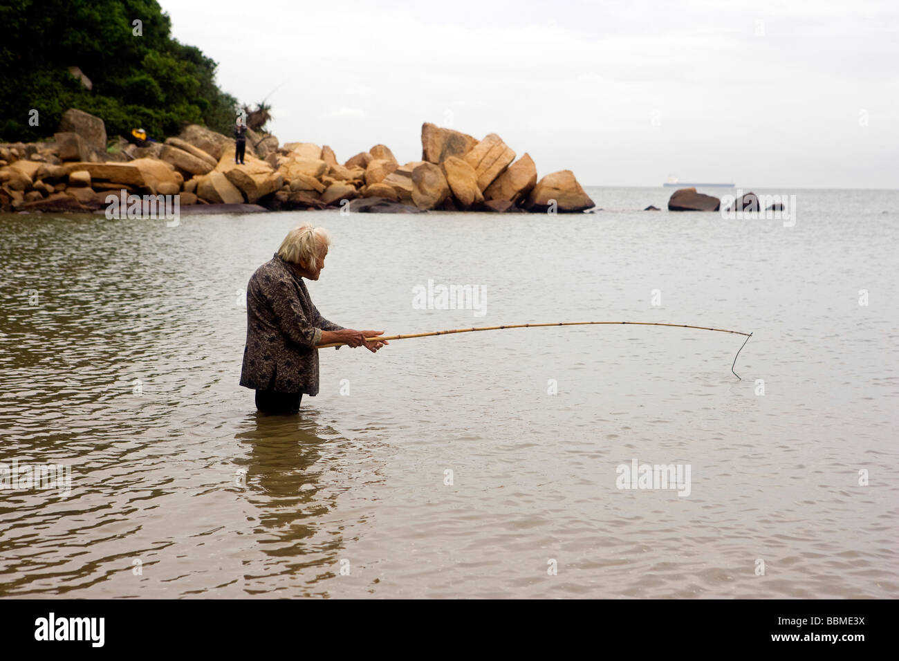 Cina, Hong Kong, l'Isola di Lantau. Una vecchia Signora sta ginocchio profondo nella pesca in mare per il suo pranzo. Foto Stock
