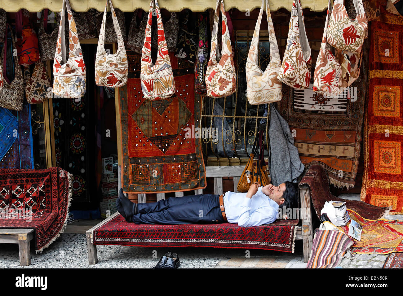 Un giovane uomo sta comodamente su un tappeto su un banco di fronte al suo negozio, utilizzando un telefono cellulare, Bazaar, Sultanahmet, Istanbul, Foto Stock