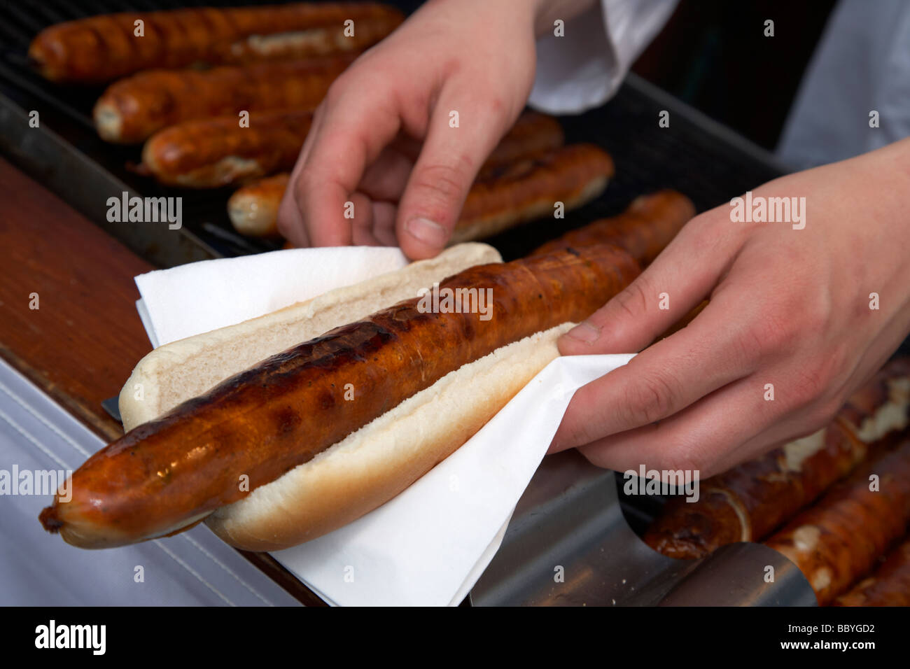 Preparazione del fornitore il bratwurst in pane bap sul tovagliolo con salsicce tedesche sul grill in vendita su un mercato all'aperto nel Regno Unito Foto Stock