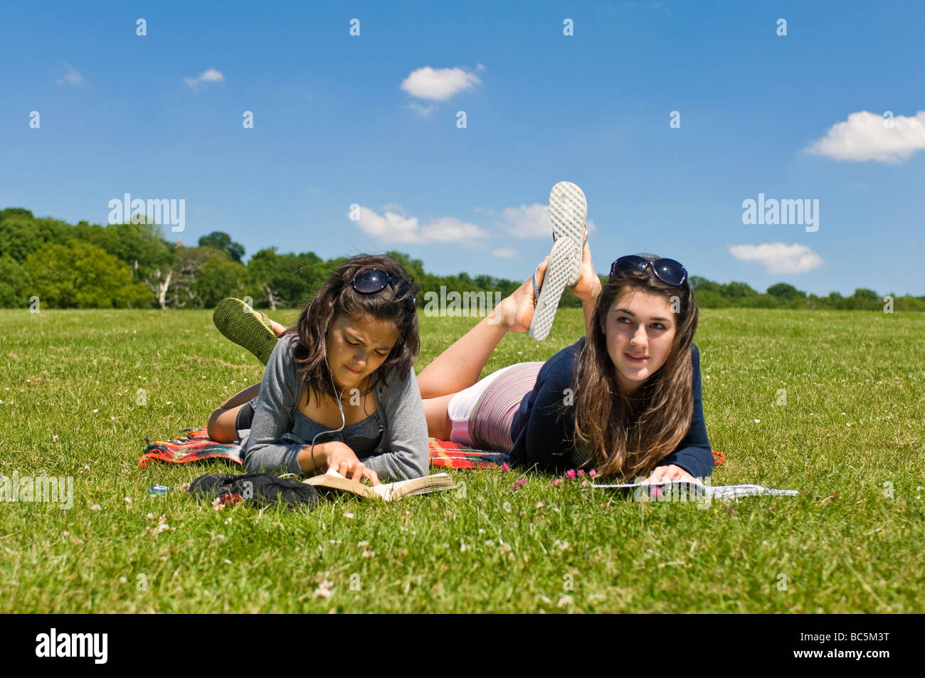 Ritratto orizzontale di due attraenti ragazze giovani sdraiati sull'erba libri di lettura e ascolto di un ipod in una giornata di sole Foto Stock