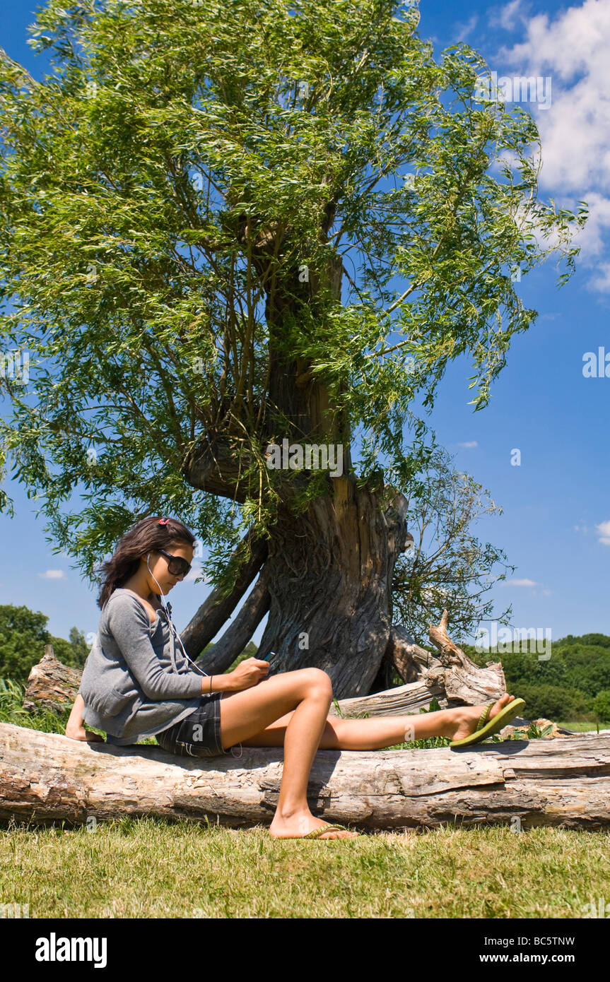 Ritratto verticale di una bella ragazza seduta su un registro l'ascolto di un ipod sotto un albero in un giorno di sole Foto Stock